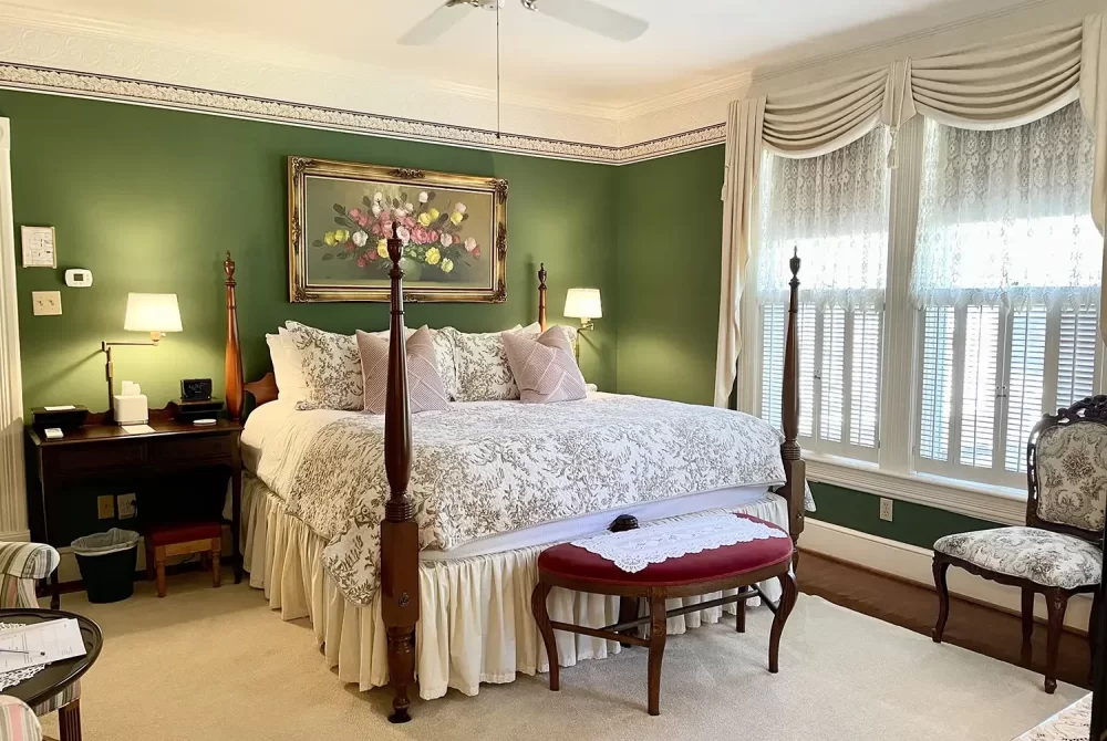 A cozy bedroom with green walls, a large bed with floral bedding, a bench at the foot, two bedside lamps, a desk, and large windows with white curtains letting in natural light—perfect for relaxing during your stay at a charming bed and breakfast in Christiansburg VA.
