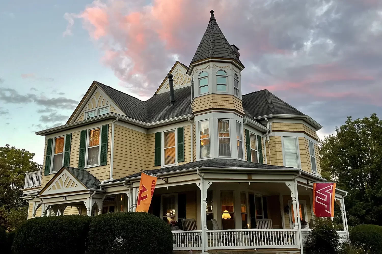 A large Victorian-style yellow house with green shutters has a wraparound porch and two flags hanging out front. Nestled in Christiansburg, VA, this charming bed and breakfast welcomes guests beneath a sky that is partly cloudy with a pinkish sunset glow.