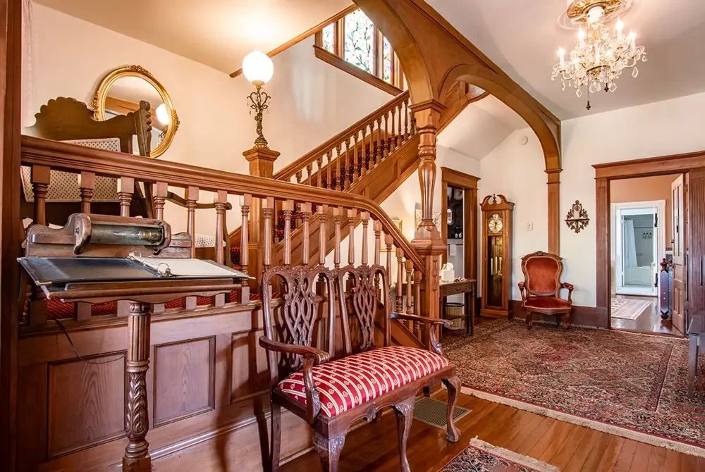 Victorian-style entryway with carved wood staircase, ornate bench, antique clock, chandelier, area rug, and upholstered armchair, featuring rich wood accents and warm lighting—the perfect welcome for guests at a charming bed and breakfast in Christiansburg, VA.