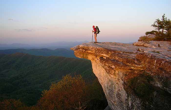 A hiker with a backpack stands on a rocky cliff edge overlooking a vast, forested valley at sunset, surrounded by distant mountains under a clear sky—perhaps inspired by the scenic landscapes near bed and breakfast Christiansburg VA.